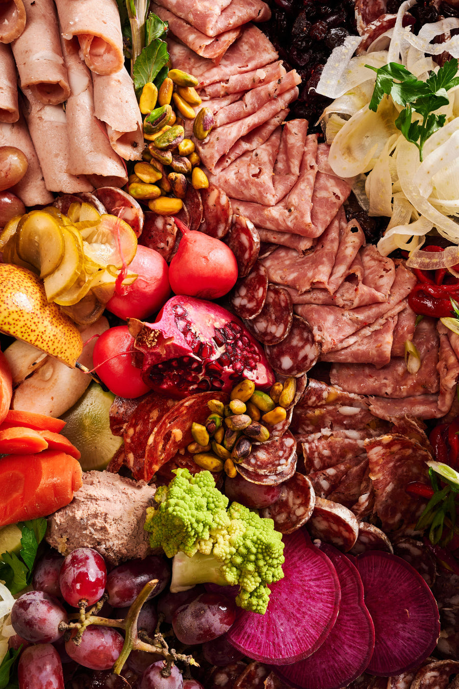 an olympia provions chefs choice board including but not limited to, ham, grapes, assortmens of salami, raisins, pistachios, potatos, carrots, olives, and pomegranates