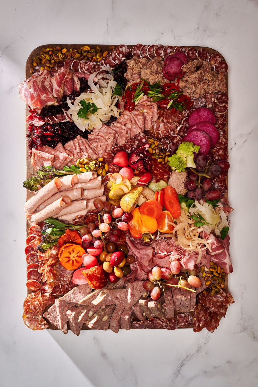 an olympia provions chefs choice board including but not limited to, ham, grapes, assortmens of salami, raisins, pistachios, potatos, carrots, olives, and pomegranates