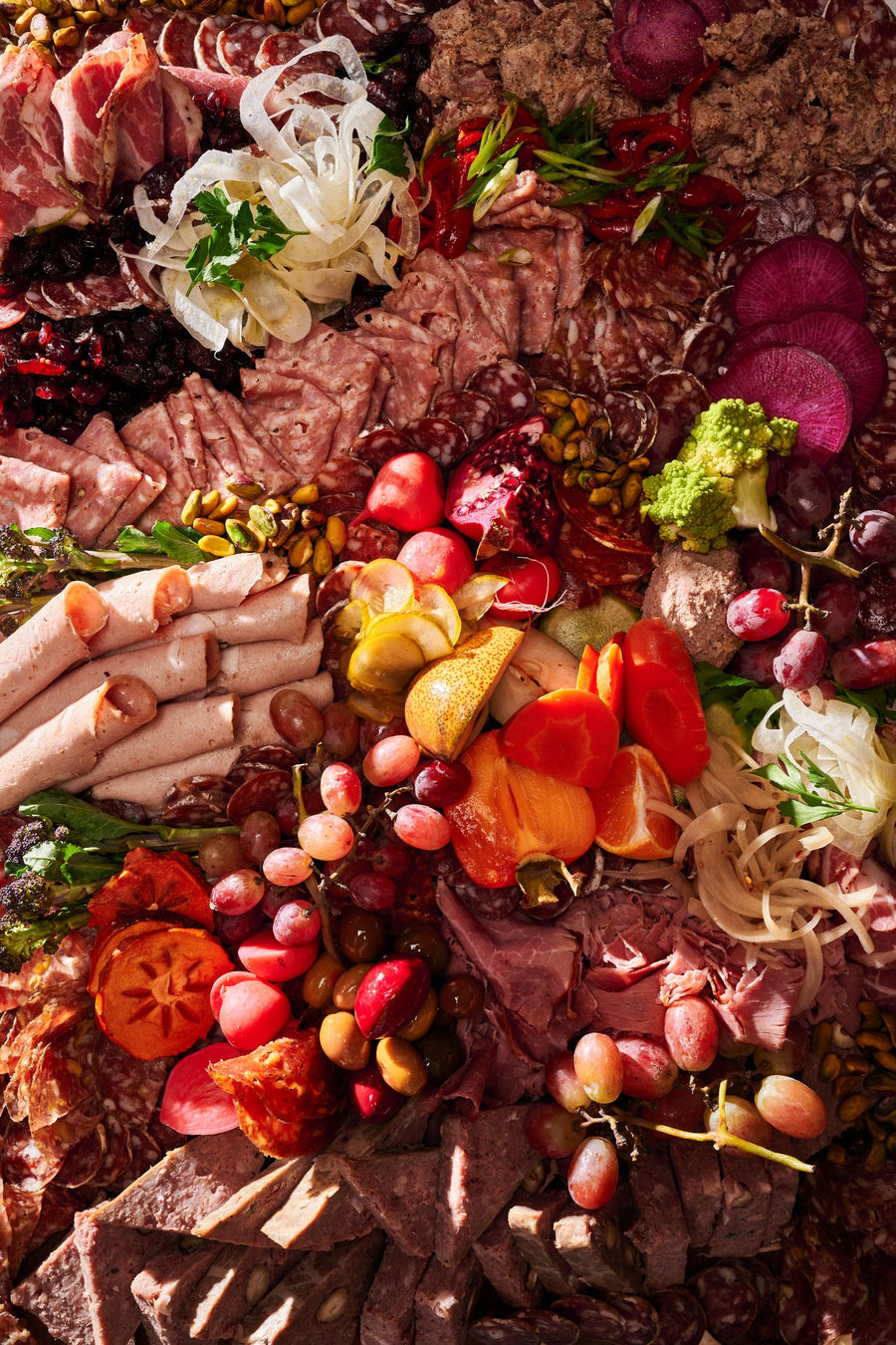 an olympia provions chefs choice board including but not limited to, ham, grapes, assortmens of salami, raisins, pistachios, potatos, carrots, olives, and pomegranates