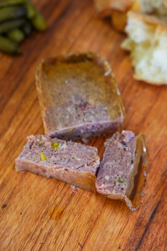 Slices of terrine on a wooden board with pickles and bread in the background