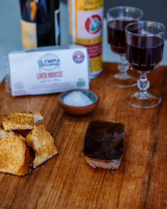 Wooden surface with toasted bread, pork liver mousse, and two glasses of red wine.