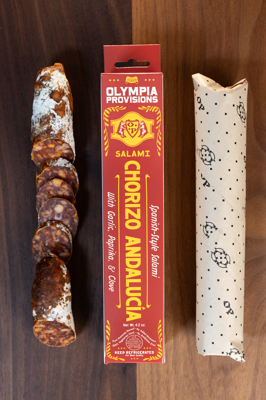 Olympia Provisions Chorizo Andalucia paprika, garlic, and clove, packaged in a red box on a wood background. A partially sliced salami sits next to the box.