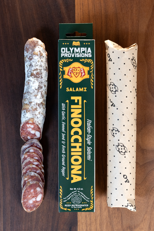 Olympia Provisions Finocchiona with garlic, black pepper, & fennel, packaged in a green box on a wood background. A partially sliced salami sits next to the box.
