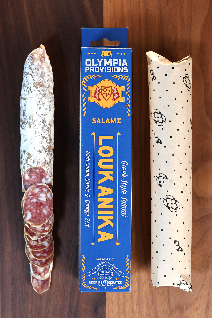 Olympia Provisions Loukanika with cumin, garlic, and orange zest, packaged in a blue box on a wood background. A partially sliced salami sits next to the box.
