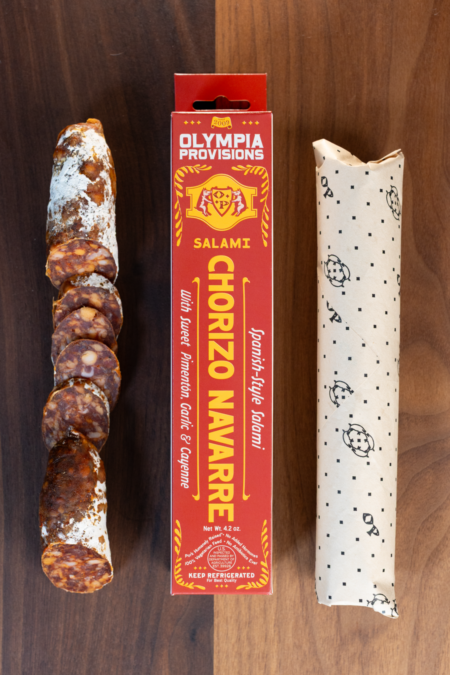 Olympia Provisions Chorizo Navarre with sweet paprika, garlic & cayenne, packaged in a red box on a wood background. A partially sliced salami sits next to the box.