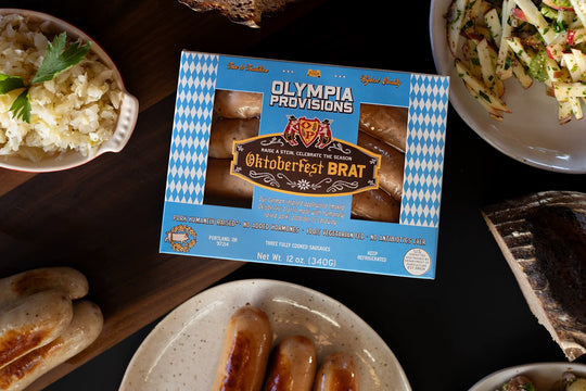 One pack of Olympia Provisions Oktoberfest Brat sausages surrounded by plates of food on a wooden table.