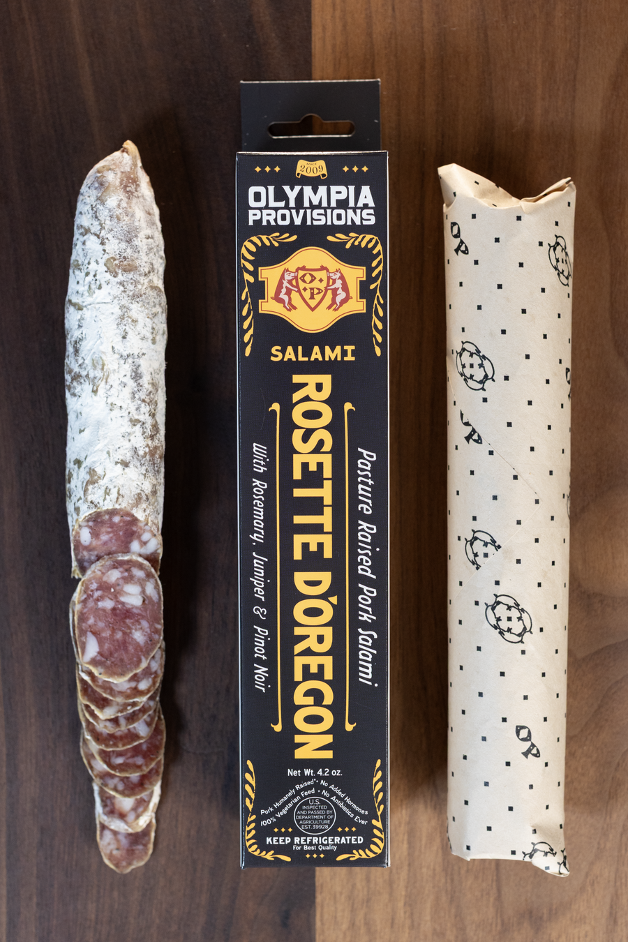 Olympia Provisions Rosette d'Oregon with rosemary, juniper, and pinot noir, packaged in a black box on a wood background. A partially sliced salami sits next to the box.