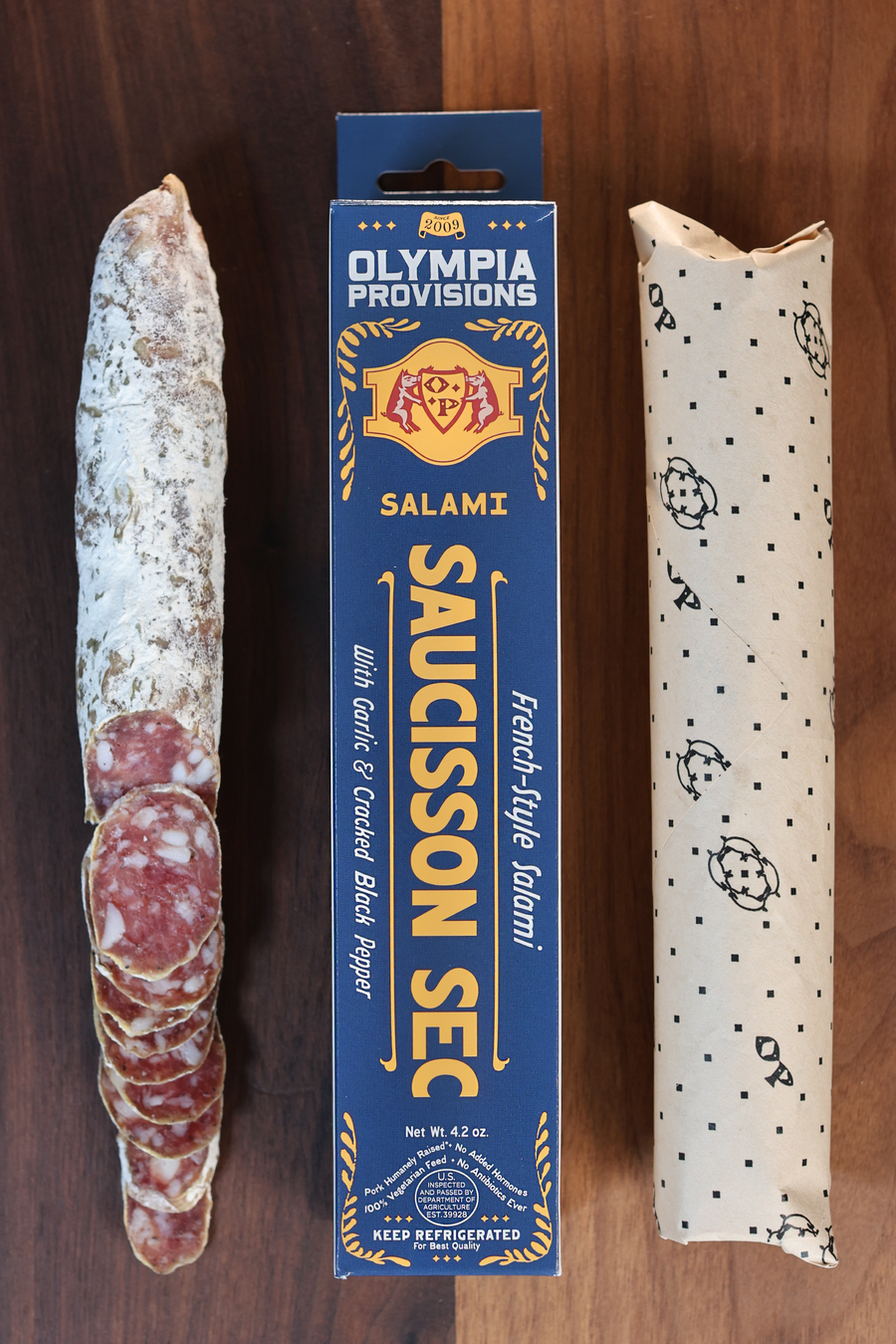 Olympia Provisions Saucisson Sec with fresh garlic and cracked black pepper, packaged in a blue box on a wood background. A partially sliced salami sits next to the box.