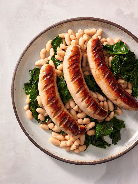A plate of grilled bratwurst served on a bed of white beans and sautéed kale. The sausages are browned with grill marks, and the dish is presented on a rustic ceramic plate against a light background.